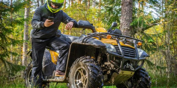 ATV rider checking phone beside parked ATV in forest, using Bloodhound Tracking to locate or recover off-road gear