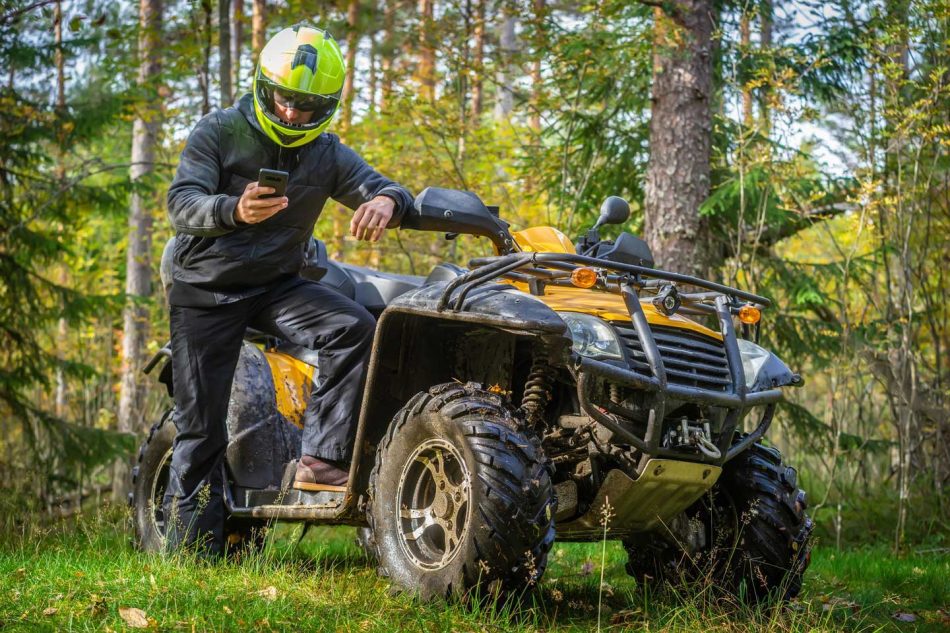 ATV rider checking phone beside parked ATV in forest, using Bloodhound Tracking to locate or recover off-road gear